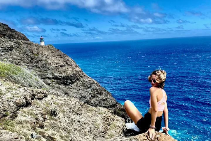 Person sitting on cliff overlooking ocean, with lighthouse in background and blue sky with clouds.