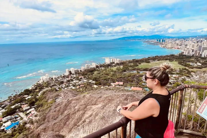 Person in athletic wear overlooks a cityscape and ocean from a hilltop railing.