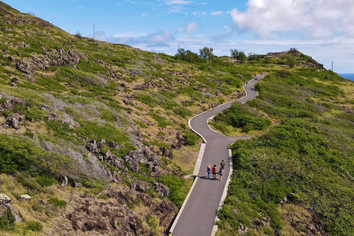 Narrow path winding through grassy hills with people walking under a blue sky.