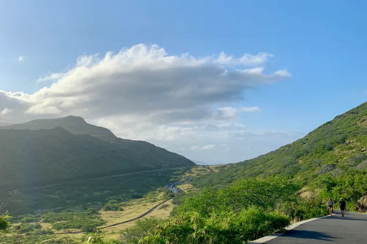 Two people walking on a road through a green valley with hills under a blue sky.