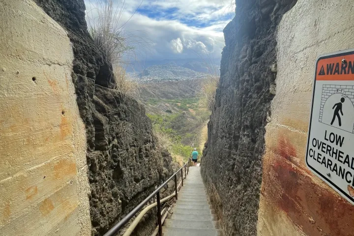 Steep staircase between rocky walls, sign warning of low clearance, cloudy sky visible at the end.